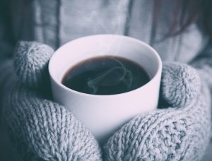 Grey mittened hands holding a white mug filled with steaming coffee. Photo by Alex on Unsplash